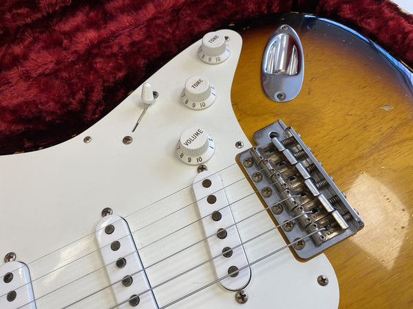 Close-up of electric guitar body showing white pickguard, tone and volume knobs, single-coil pickups, strings, and metal bridge on sunburst finish guitar in red plush case interior