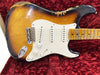 Vintage sunburst electric guitar with white pickguard, three single-coil pickups, and maple fretboard showing heavy wear and finish checking, resting on red plush guitar case lining