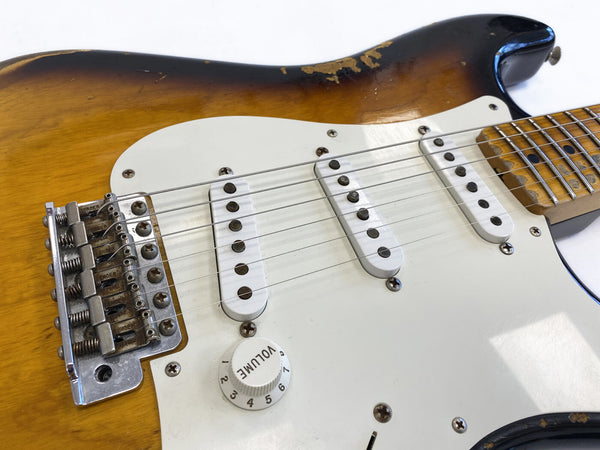 Close-up of vintage sunburst electric guitar body showing white pickguard, three single-coil pickups, volume knob, and worn maple neck with Fender branded bridge saddles