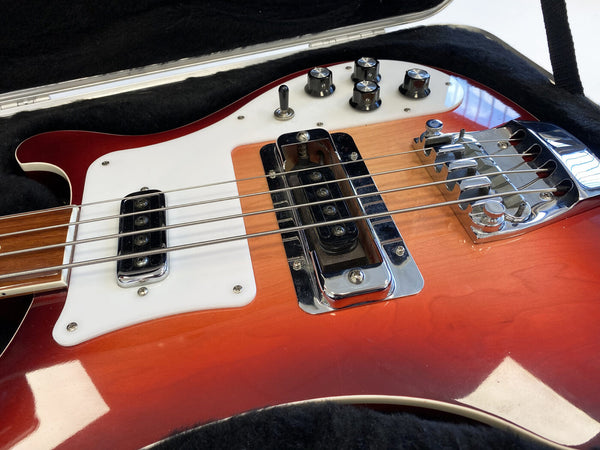 Close-up of vintage red electric bass guitar with white pickguard, chrome pickups, and black control knobs inside a black padded guitar case