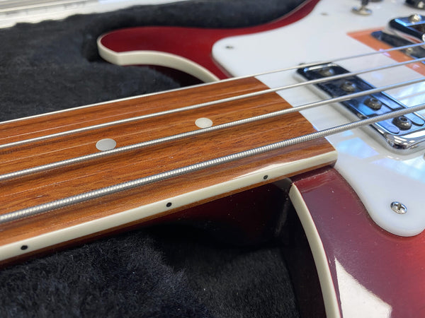 Close-up of red electric bass guitar neck, focusing on rosewood fretboard with silver strings and white dot inlays, lying on black fabric case interior.