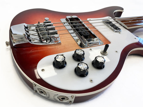 Close-up of a vintage electric bass guitar body with sunburst finish, featuring chrome bridge, pickup covers, black control knobs labeled for volume, tone, treble, and bass, and input jacks on a white pickguard.