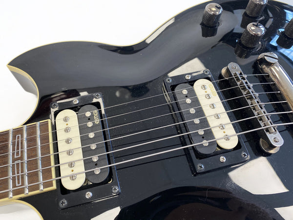 Close-up of black electric guitar body featuring dual EMG humbucker pickups, metal bridge, and control knobs with cream binding and rosewood fretboard