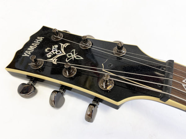 Close-up of Yamaha guitar headstock with black finish, tuning pegs, and decorative inlay on white background