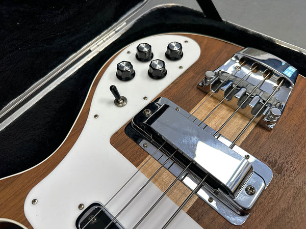 Close-up of electric bass guitar body showing chrome bridge, control knobs, toggle switch, and wooden finish in protective case