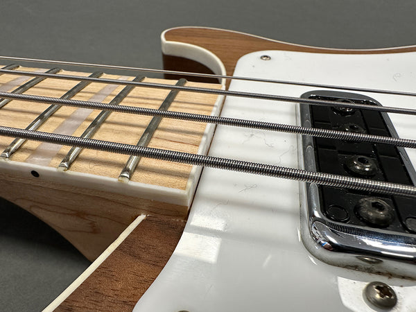 Close-up of electric bass guitar neck and pickups showing metal frets, thick metal strings, maple fretboard, and white pickguard with wood body edges