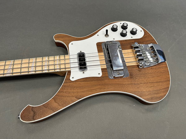 Close-up of a natural wood finish electric bass guitar with white pickguard, chrome bridge and pickups, black control knobs, and maple neck with block inlays on a gray background