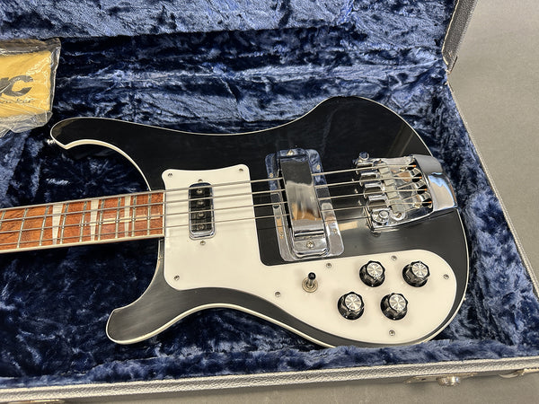 Black electric bass guitar with white pickguard and control knobs resting inside a blue plush lined hard case