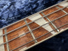 Close-up image of bass guitar neck showing metal strings, wooden fretboard with silver triangular inlays, and metal frets on a blue plush surface.
