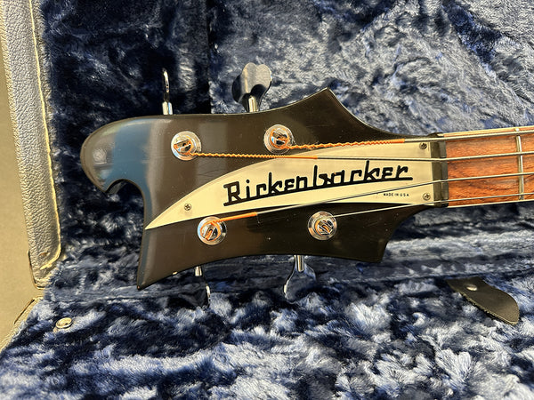 Close-up of black Rickenbacker bass guitar headstock with tuning pegs, Rickenbacker logo, and strings on plush blue guitar case interior.