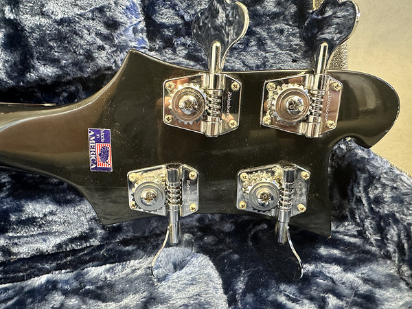 Close-up of black bass guitar headstock with four chrome tuning pegs and Made in America sticker resting on blue plush fabric case interior