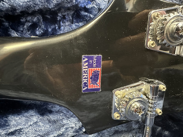 Close-up of black guitar headstock with chrome tuning machines and a "Made in America" sticker on a blue fabric background