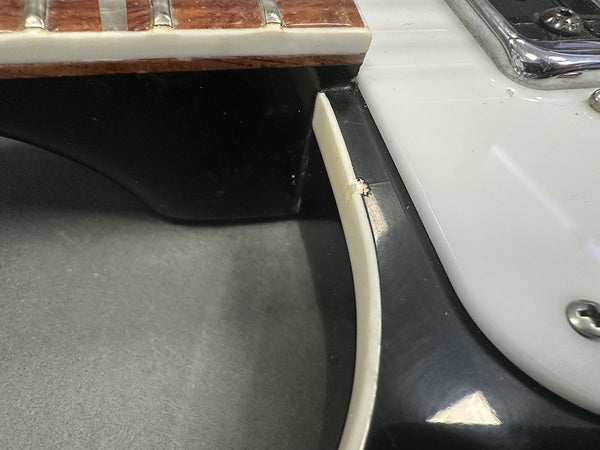 Close-up detail of guitar neck joint area showing nut and fretboard binding with minor wear on vintage electric guitar body edge near white pickguard and black finish