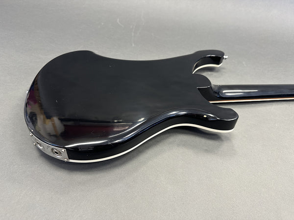 Black electric guitar body and neck with white binding on gray surface background, rear view showing guitar input jack and contour details.
