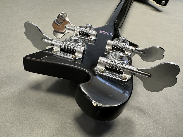 Close-up of black bass guitar headstock showing chrome tuning pegs and minor wear on paint edges
