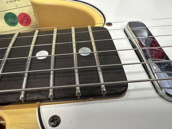 Close-up of electric guitar fretboard with metal frets, mother-of-pearl dot inlays, steel strings, and chrome pickup on white body.