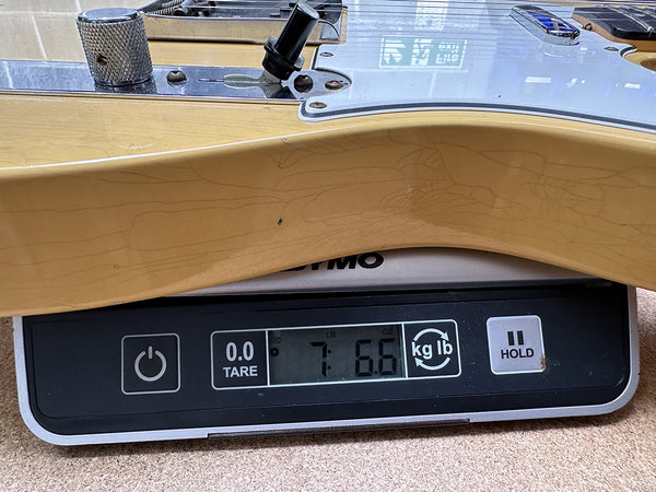 Close-up of a light-colored electric guitar body on a digital scale showing a weight of 7 pounds 6.6 ounces, with visible hardware and control knobs.