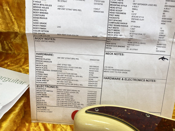Close-up of a guitar specification sheet detailing hardware, electronics, body notes, and neck notes for a 60 Strat model with aged vintage white finish, alongside the back of a vintage-style guitar with a tortoiseshell pickguard, placed on a yellow velvet surface.
