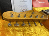 Close-up of Fender Stratocaster guitar headstock with vintage tuning pegs and original decal, displayed on yellow plush lining with Fender certificate and owner's manual in background.