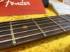 Close-up of Fender guitar rosewood fretboard with strings and fret markers on yellow fabric background and Fender owner's manual in the background.