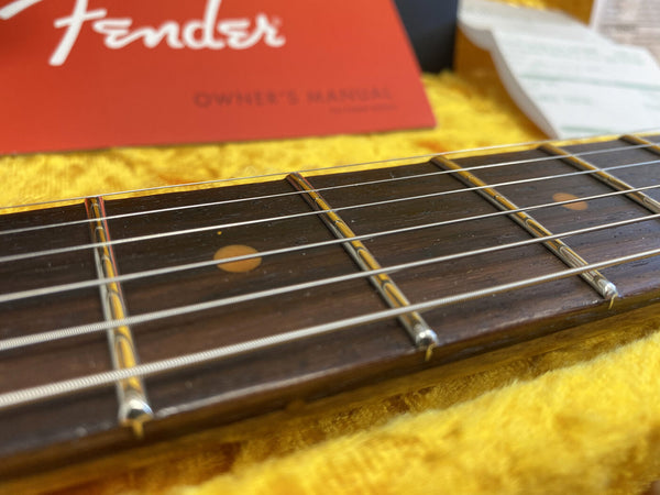 Close-up of Fender guitar rosewood fretboard with strings and fret markers on yellow fabric background and Fender owner's manual in the background.