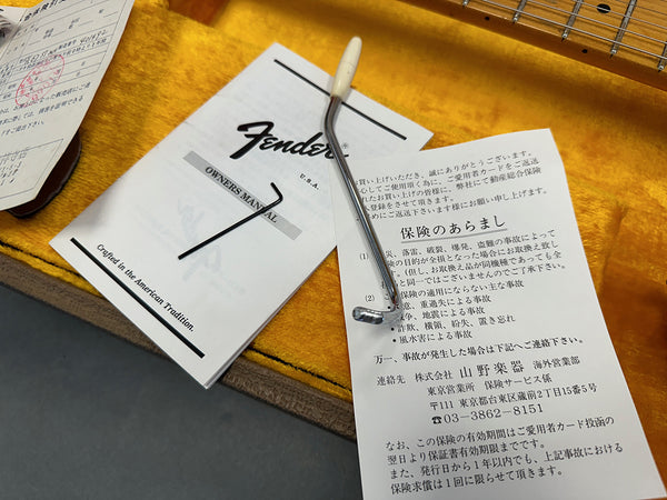 Fender owner's manual with tremolo arm and hex wrench on yellow guitar case lining next to a guitar neck