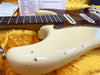 Close-up of a vintage cream electric guitar body with visible wear and paint chips, showing tortoiseshell pickguard, white single-coil pickups, and part of the rosewood fretboard, resting on a yellow plush case interior with paperwork in the background.