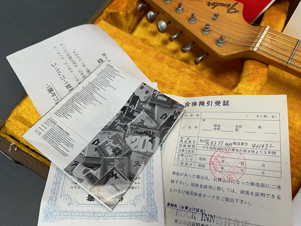 Fender electric guitar headstock in vintage case with yellow plush interior and accompanying Japanese warranty papers and Fender string packaging.