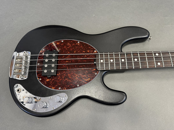 Black electric bass guitar with tortoiseshell pickguard and chrome control knobs on gray background