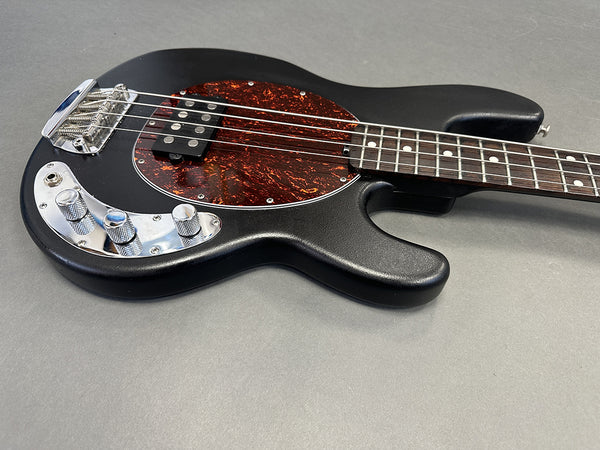 Black electric bass guitar with tortoiseshell pickguard and chrome control knobs on gray background