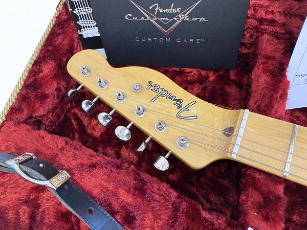 Close-up of Fender Custom Shop guitar headstock with tuning pegs inside red plush-lined guitar case with Fender Custom Shop documentation and black leather strap.