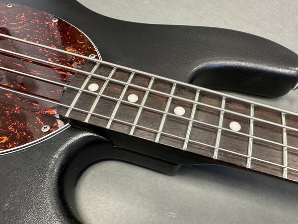 Close-up of guitar fretboard with metal frets and white dot inlays, black textured guitar body, and tortoiseshell pickguard with metal screws.