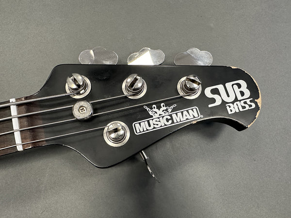 Close-up of Music Man SUB Bass guitar headstock with tuning pegs and worn edges on black finish
