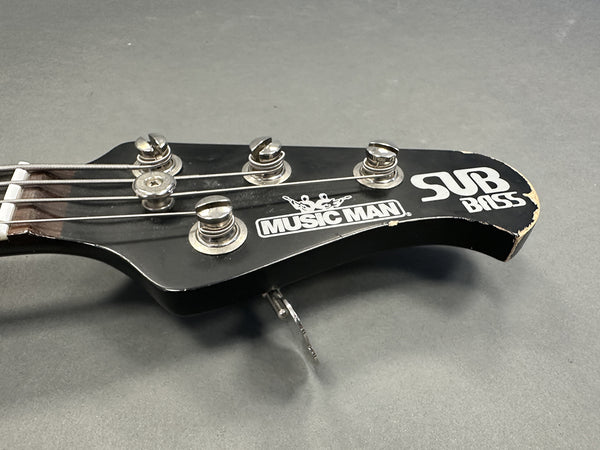 Close-up of black Music Man Sub Bass guitar headstock with tuning pegs and worn edges on gray background