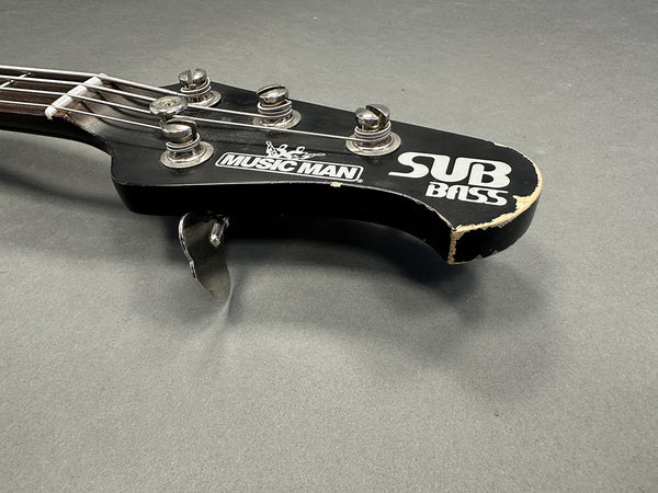 Close-up of black Music Man SUB Bass guitar headstock with tuning pegs and worn edges on gray background