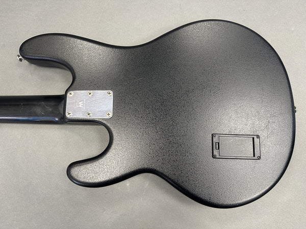 Black electric guitar back body with textured finish and neck plate screws visible, showing battery compartment door on gray surface