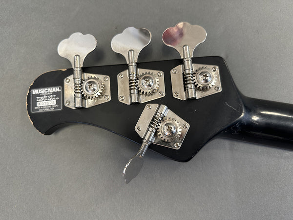 Close-up of black Music Man bass guitar headstock with four silver tuning pegs and Ernie Ball brand label made in San Luis Obispo, California USA