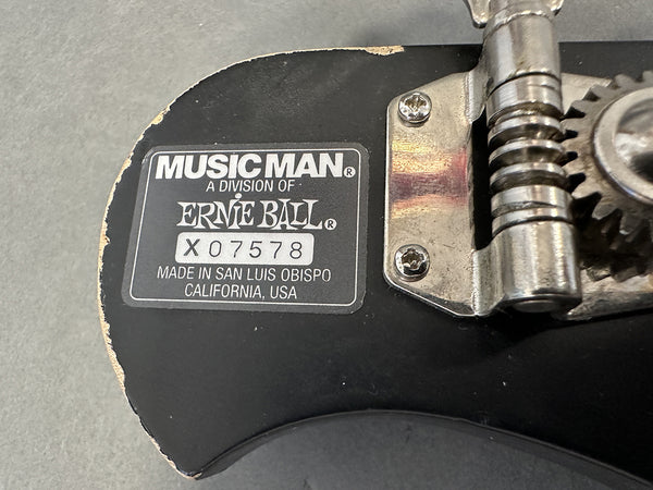 Close-up of Music Man Ernie Ball serial number X07578 label and tuning gear on guitar headstock showing manufacturing details from San Luis Obispo, California