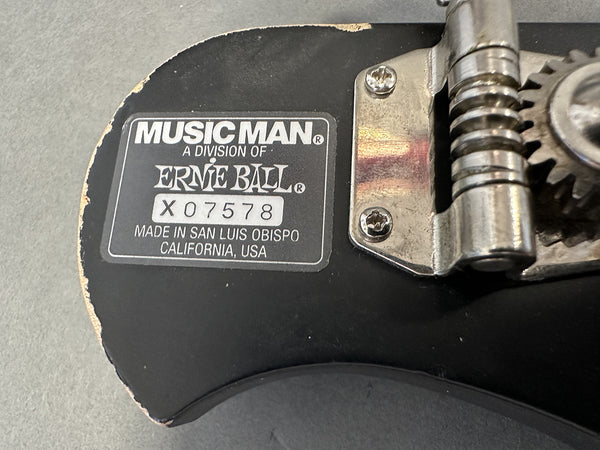 Close-up of Music Man Ernie Ball serial number X07578 label and tuning mechanism on black guitar body made in San Luis Obispo California USA with wear on edge