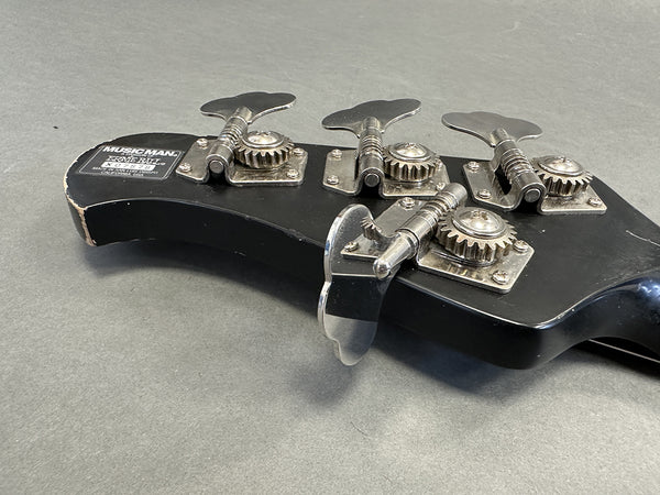 Close-up of Music Man electric bass guitar headstock with chrome tuning pegs and Ernie Ball label on black finish