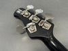 Close-up of black Music Man bass guitar headstock with four silver tuning pegs and tuning gears on gray background