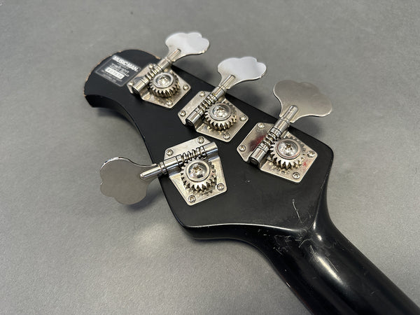 Close-up of black Music Man bass guitar headstock with four silver tuning pegs and tuning gears on gray background