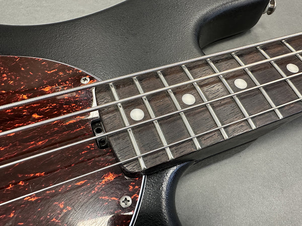 Close-up of black electric bass guitar fretboard with silver frets, metal strings, and tortoiseshell pickguard on gray surface