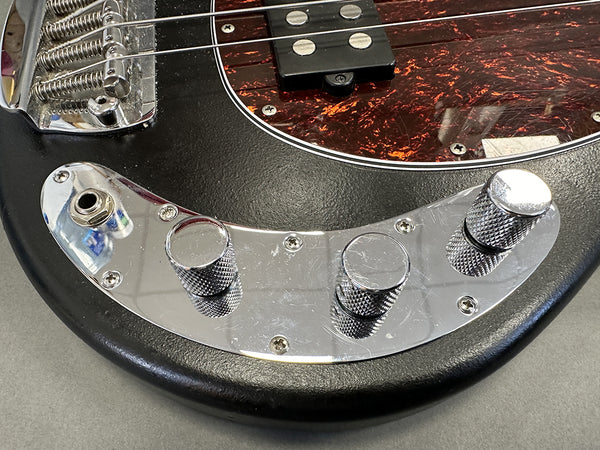 Close-up of black electric bass guitar with chrome control plate and knobs, tortoiseshell pickguard, and metal bridge on gray background