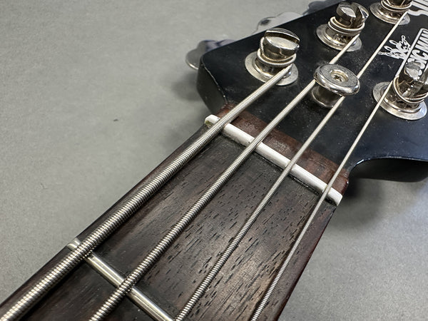 Close-up of a bass guitar headstock and fretboard with metal strings and tuning pegs on a gray background