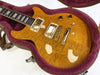 Electric guitar with quilted maple top in honey amber finish, two humbucker pickups, three control knobs, and toggle switch, resting in purple plush-lined brown hard case