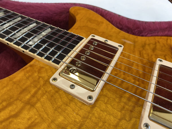 Close-up view of honey-colored quilted maple electric guitar body with cream pickup rings and humbucker pickups, detailed fretboard with pearl inlays, and metal strings on a purple velvet guitar case background.