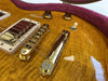Close-up of amber quilted maple electric guitar body with gold bridge, pickup, control knobs, and toggle switch in plush pink guitar case lining