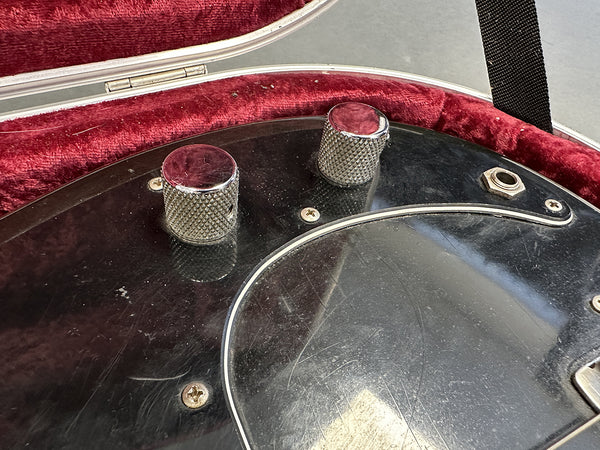 Close-up of black electric guitar control knobs and input jack on scratched pickguard inside red velvet-lined guitar case