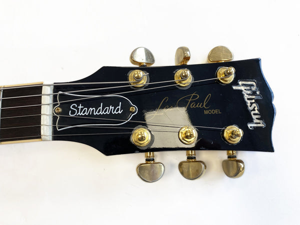 Close-up of black Gibson Les Paul Standard guitar headstock with gold tuning pegs and white logo on a white background